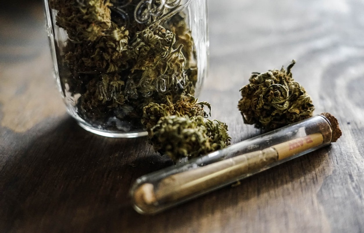 Cannabis flower in a glass jar beside a pre-roll on a wooden surface